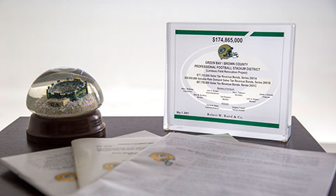 Photograph featuring: a Lambeau Field snowglobe, placard regarding Lambeau Field Renovation Project deal, copies of the final official statement regarding the project deal with Baird. 