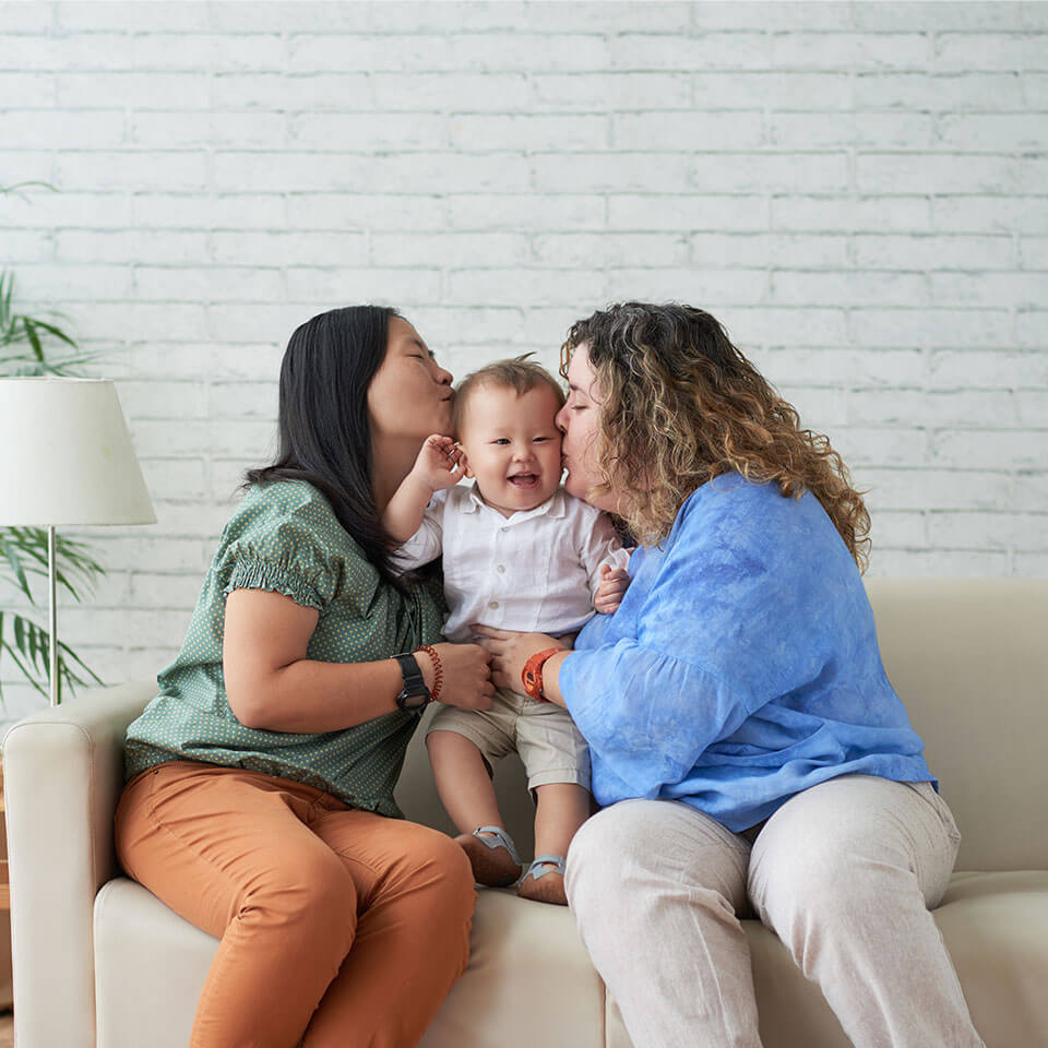 Same-sex couple kissing their smiling toddler on both cheeks. Asian and Caucasian ethnicity, dark brown and curly, blonder hair. Living room with lots of natural light.