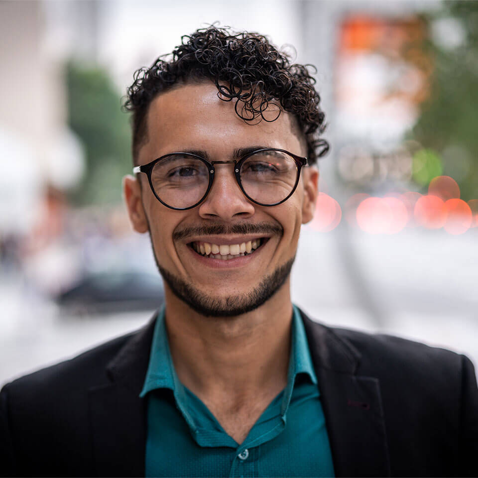 Portrait of hispanic male with round, black glasses, curly, dark brown hair and beard wearing a teal button down and black blazer with blurred street in the background.