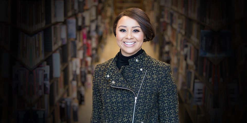 Photo of Devon Norwood, brunette hair pulled back in bun and wearing dark tweed blazer, standing in front of library aisle.