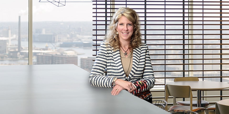 Photo of Kim Haws Falasco, medium length blonde hair and wearing black and white striped blazer, standing in Baird break room.