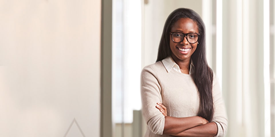 Photo of Nina Essandoh, long bruntter hair and wearing oatmeal colored sweater and glasses, standing in office setting.