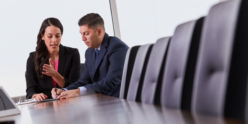 Photograph of Andi Mills, long brunette hair wearing coral shirt and black blazer, and Nick Gomez, short crew cut wearing navu suit, sitting at conference table going over documents.